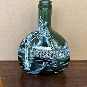 May include: A dark green glass bottle with a white painted scene of a stone building with a water wheel, trees, and a fence. The bottle has a round body and a narrow neck.
