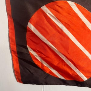 May include: A silk scarf with a bold geometric design. The scarf features a large orange circle with white stripes against a black background. The edges are trimmed with orange.