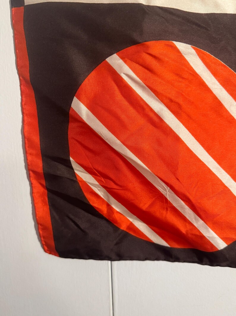 May include: A silk scarf with a bold geometric design. The scarf features a large orange circle with white stripes against a black background. The edges are trimmed with orange.