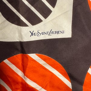 May include: A silk scarf with a repeating pattern of brown and orange circles with white stripes. The scarf has the text "Yves Saint Laurent" in navy blue.