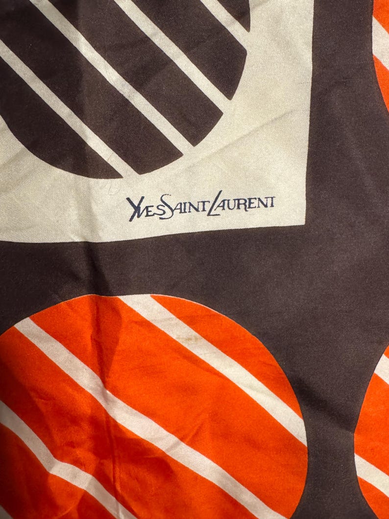 May include: A silk scarf with a repeating pattern of brown and orange circles with white stripes. The scarf has the text "Yves Saint Laurent" in navy blue.