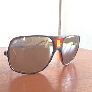 May include: Brown-tinted sunglasses with a black frame and red and yellow accents. The sunglasses have a retro aviator style with a thick frame and a bridge. The lenses are a dark brown color, and the arms are a mix of yellow and black.
