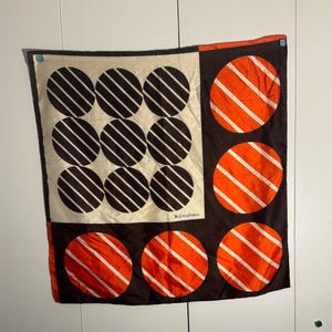 May include: A square silk scarf with a geometric design. The scarf features a cream-colored square with nine black circles, each with white diagonal stripes. The other sections are brown with orange circles and white stripes. The scarf has a designer's signature.