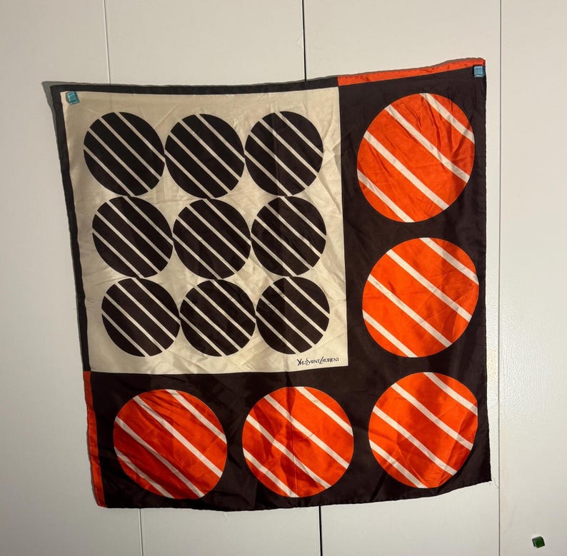 May include: A square silk scarf with a geometric design. The scarf features a cream-colored square with nine black circles, each with white diagonal stripes. The other sections are brown with orange circles and white stripes. The scarf has a designer's signature.