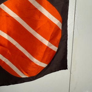 May include: A dark brown scarf with a large orange circle design featuring white diagonal stripes. The scarf is made of a silky material and has a smooth texture. The design is centered on the scarf.
