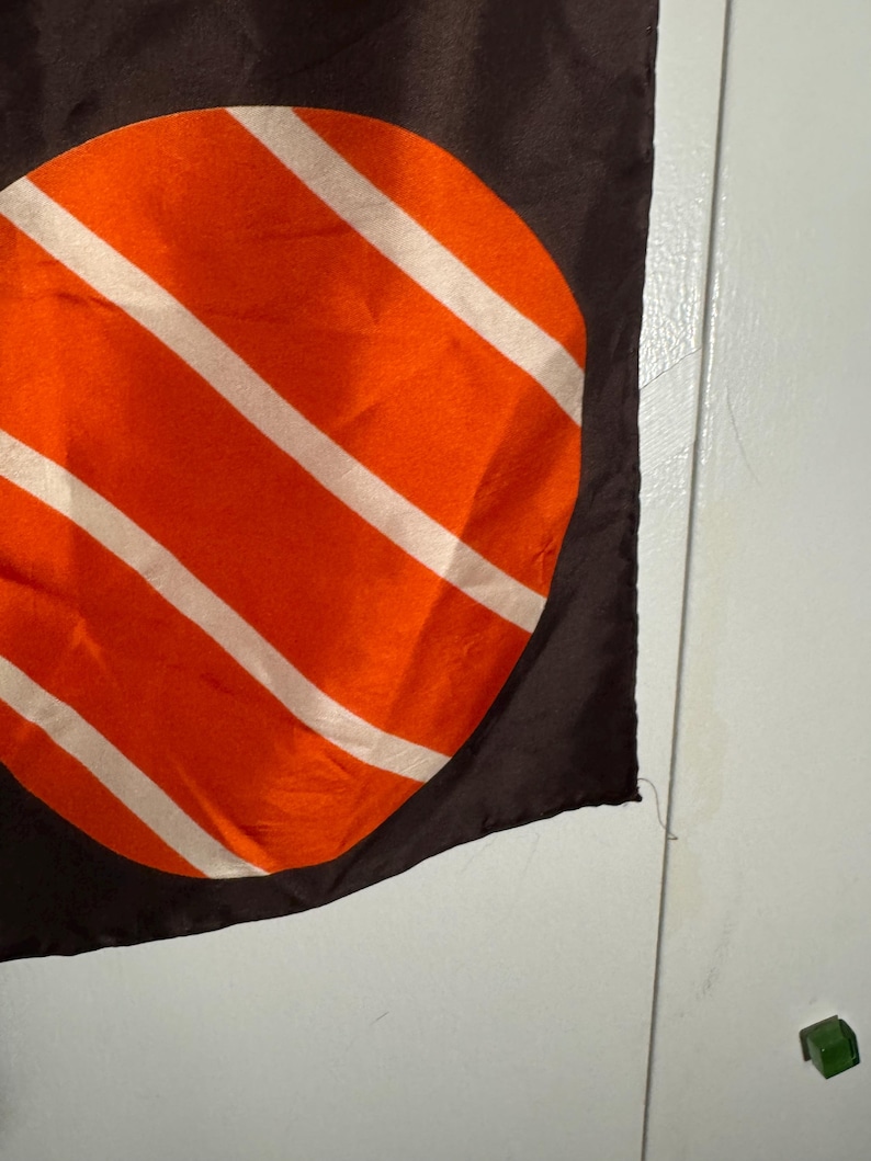 May include: A dark brown scarf with a large orange circle design featuring white diagonal stripes. The scarf is made of a silky material and has a smooth texture. The design is centered on the scarf.