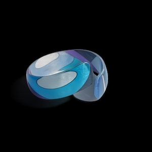 May include: A modern, abstract bracelet with a swirling design. The bracelet features shades of blue, white, and purple. The transparent material allows light to pass through, creating a play of shadows. The bracelet is a unique piece of jewelry.