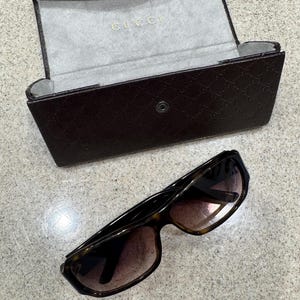 May include: A pair of brown tortoise shell sunglasses with gradient lenses positioned below a dark brown hard-shell glasses case. The case features a repeating embossed logo pattern on the exterior and the word GUCCI printed in gold inside.