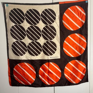 May include: A square silk scarf with a geometric design. The scarf features a cream-colored section with nine black circles, each with white diagonal stripes. The other sections are brown with orange circles and white stripes. The Yves Saint Laurent logo is visible.