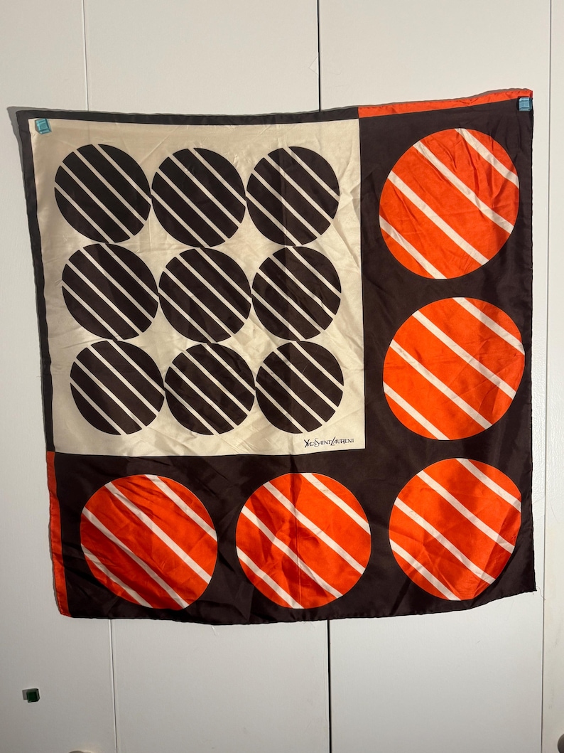 May include: A square silk scarf with a geometric design. The scarf features a cream-colored section with nine black circles, each with white diagonal stripes. The other sections are brown with orange circles and white stripes. The Yves Saint Laurent logo is visible.