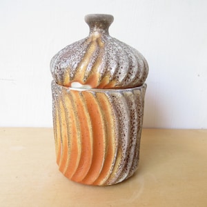 May include: A ceramic jar with a lid. The jar is cylindrical with vertical, curved ridges in shades of orange, brown, and gray. The lid has a similar design and a small, rounded knob. The jar is on a light-colored surface.