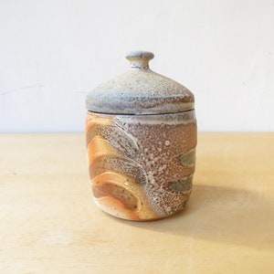 以下が含まれることがあります： 蓋付きの陶器の壺。壺は円筒形で、丸い底と小さなつまみのある蓋が付いています。壺は茶色、オレンジ色、白の渦巻き模様です。蓋はグレーで、質感があります。