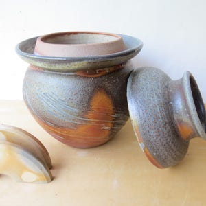 Puede incluir: Juego de cerámica artesanal con un jarrón grande y redondo con tapa, un recipiente interior más pequeño y dos piezas decorativas en forma de media luna. El jarrón es marrón, naranja y azul con una textura moteada.
