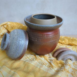 Puede incluir: Una mantequera de cerámica con una base de color marrón rojizo intenso y una tapa texturizada de color gris claro. El recipiente principal presenta un borde ancho y un interior liso. Dos pequeñas piezas de cerámica curvas en tonos gris y marrón.