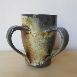 May include: A handcrafted ceramic vase with two curved handles. The vase features a dark, mottled glaze with shades of brown, gold, and black. The handles are a darker shade of gray. The vase has a wide opening and a tapered base.