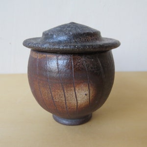 May include: A handcrafted, ceramic jar with a lid. The jar is a warm brown color with vertical, textured lines. The lid is a darker, charcoal gray. The jar has a rounded body and a small, pedestal base.