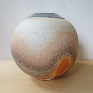 Puede incluir: Un jarrón redondo de cerámica con un esmalte blanco moteado y un esmalte marrón y naranja en la parte inferior. El jarrón tiene un borde gris oscuro.