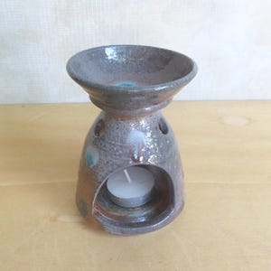 May include: A ceramic essential oil diffuser with a tealight candle inside. The diffuser is a mottled brown and gray with teal and brown accents. It has a bowl-shaped top and a round base with air holes.