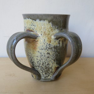 May include: A ceramic mug with two handles, featuring a mottled glaze in shades of gray, yellow, and brown.