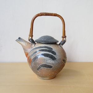 Peut inclure: Une théière en céramique avec un corps rond et une anse en bambou tressé. La théière a un couvercle gris et un bec verseur. Le corps est décoré de lignes courbes noires et grises sur un fond beige et orange. La théière est posée sur une surface marron clair.