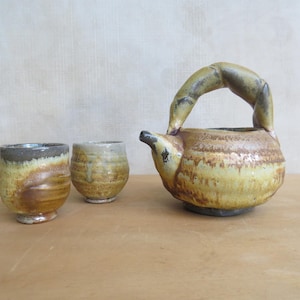 Puede incluir: Un juego de té de cerámica hecho a mano con una tetera con asa curva y dos tazas a juego. El juego tiene una estética rústica y terrosa con una mezcla de tonos marrones, amarillos y grises. La tetera mide aproximadamente 15 cm de alto.
