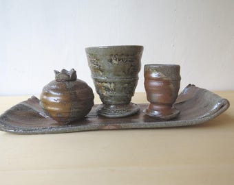 Op de afbeelding: Een handgemaakte keramische set met een granaatappelvormige container, een grote beker en een kleinere beker, allemaal rustend op een lange, rechthoekige tray. De stukken zijn geglazuurd in aardetinten van bruin, beige en grijs.