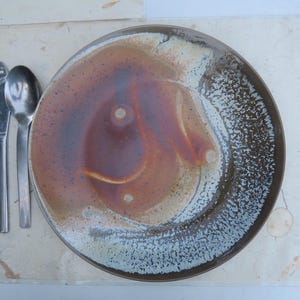 Puede incluir: Un plato de cerámica con un esmalte marrón y beige, acompañado de un tenedor, un cuchillo y una cuchara. El plato tiene un borde moteado y un diseño orgánico. La cubertería es de acero inoxidable.