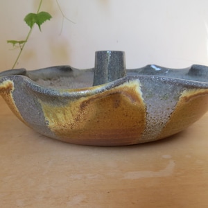 May include: A handmade ceramic bowl with a central cylindrical feature. The bowl has a wavy rim and is colored with shades of gray, brown, and yellow. This unique piece is suitable for decorative or functional use.