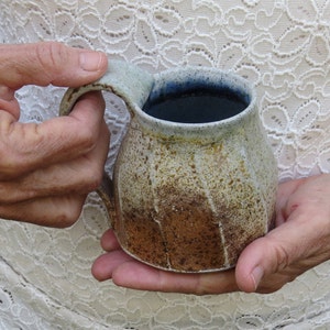 Puede incluir: Una taza de cerámica hecha a mano con un diseño moteado y terroso. La taza presenta una base marrón que se desvanece en un borde beige y azul claro. La taza está sostenida por una persona.