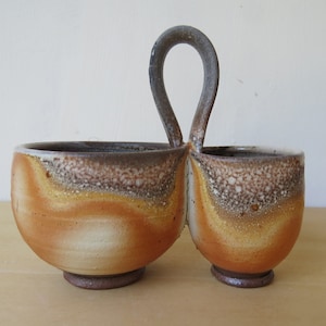May include: A ceramic serving dish with two bowls and a handle. The bowls are brown, white, and yellow with a speckled glaze. The handle is brown and is shaped like a loop.