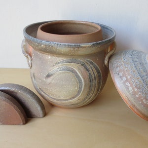 May include: A ceramic pot with a lid and two small ceramic supports. The pot is decorated with a swirling pattern in shades of brown, grey, and white. The lid is also decorated with a similar pattern.
