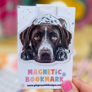 May include: A white magnetic bookmark with a brown and white dog's face on it. The bookmark says "MAGNETIC BOOKMARK" and "www.gingerpatchdesigns.com".