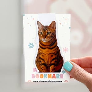 May include: A decorative bookmark featuring a Bengal cat illustration. The bookmark has a white background with a colorful floral design and the word "BOOKMARK" in pastel colors. The website address "www.elmerratchdesigns.com" is printed at the bottom.