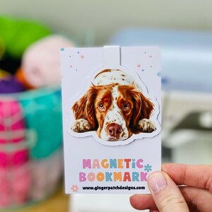 May include: A white and brown dog with a black nose is featured on a white magnetic bookmark. The bookmark has pink, blue, and light blue polka dots and the text "MAGNETIC BOOKMARK" in colorful letters. The text "www.gingerpatchdesigns.com" is printed at the bottom of the bookmark.