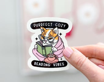 May include: A white sticker with a cartoon cat reading a book, wearing glasses and wrapped in a pink blanket. The sticker has the text "PURRFECT COZY" and "READING VIBES". A pink teacup sits next to the cat.