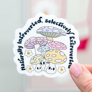 May include: A die-cut sticker featuring a cluster of cartoon mushrooms in pastel colors. The sticker has the text "Naturally Introverted, Selectively Extroverted" in a circular design around the mushrooms. The mushrooms have smiling faces and are surrounded by small flower illustrations.