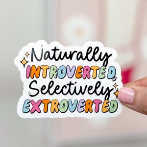 May include: A white sticker with the words "Naturally Introverted, Selectively Extroverted" in a colorful, bubble-style font. The sticker has a black outline and is held by a person.
