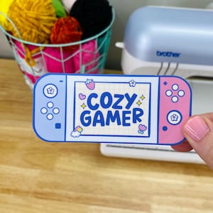 May include: A pink and blue sticker with a Nintendo Switch design. The screen of the sticker says "Cozy Gamer" in blue text with a pink heart and stars.