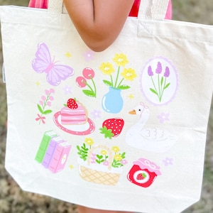 May include: Cream-colored tote bag featuring a whimsical design of a butterfly, flowers, cake, strawberries, a swan, and books. The illustrations are in soft pastel colors, creating a charming and playful aesthetic. The bag is held by a person.