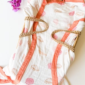 May include: A white muslin baby blanket with a coral pink border and a seashell print. The blanket is folded and placed in a woven basket.
