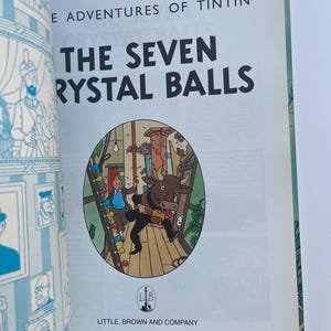 May include: Open book featuring the cover of "The Seven Crystal Balls" by Herg&eacute;. The title is in large black letters. A circular illustration depicts a scene with a bull and a character. The publisher's name and city are at the bottom.