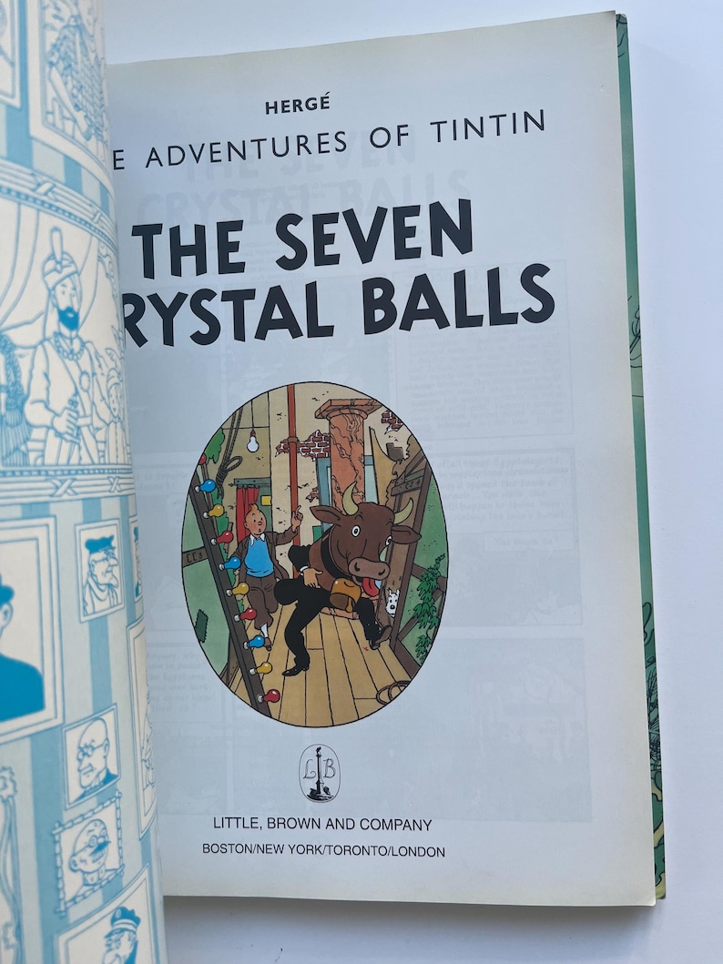 May include: Open book featuring the cover of "The Seven Crystal Balls" by Herg&eacute;. The title is in large black letters. A circular illustration depicts a scene with a bull and a character. The publisher's name and city are at the bottom.