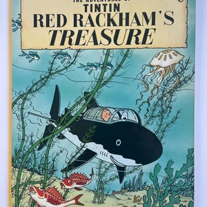 May include: A vintage book cover for "The Adventures of Tintin: Red Rackham's Treasure." The cover features a shark-shaped submarine with Tintin and Snowy inside, surrounded by marine life. The title is in bold red letters.