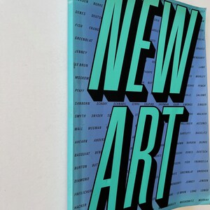 K&ouml;nnte beinhalten: Ein Buch mit dem Titel "NEW ART" mit gro&szlig;en t&uuml;rkisfarbenen Buchstaben und schwarzen Schatten. Der Einband des Buches ist hellblau und zeigt eine Liste von Namen in wei&szlig;em Text. Das Buch wird schr&auml;g dargestellt.