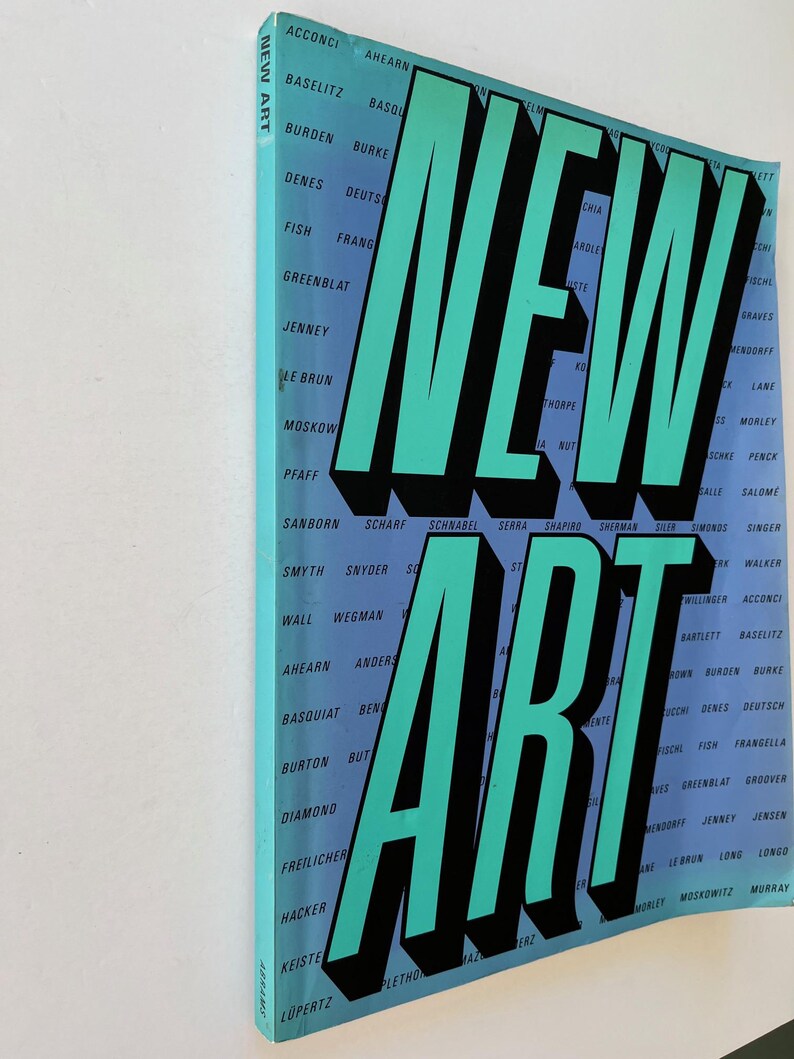 K&ouml;nnte beinhalten: Ein Buch mit dem Titel "NEW ART" mit gro&szlig;en t&uuml;rkisfarbenen Buchstaben und schwarzen Schatten. Der Einband des Buches ist hellblau und zeigt eine Liste von Namen in wei&szlig;em Text. Das Buch wird schr&auml;g dargestellt.