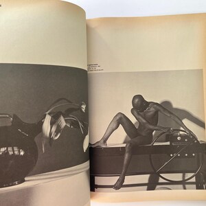 K&ouml;nnte beinhalten: Schwarz-Wei&szlig;-Fotografie aus einem Buch. Links eine dunkle Kanne mit Henkel und Bl&auml;ttern. Rechts sitzt eine nackte Person auf einem dunklen Objekt mit einem kreisf&ouml;rmigen Rad.