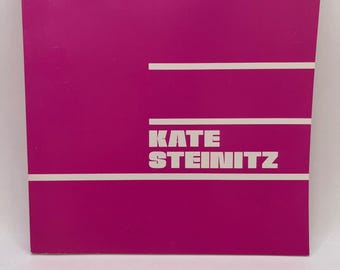 May include: A pink pamphlet with white text that reads "KATE STEINITZ".