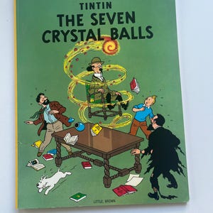 May include: A hardcover book titled "The Adventures of Tintin: The Seven Crystal Balls" with a green cover. The cover features a colorful illustration of characters and objects, including a table, books, and a dog.