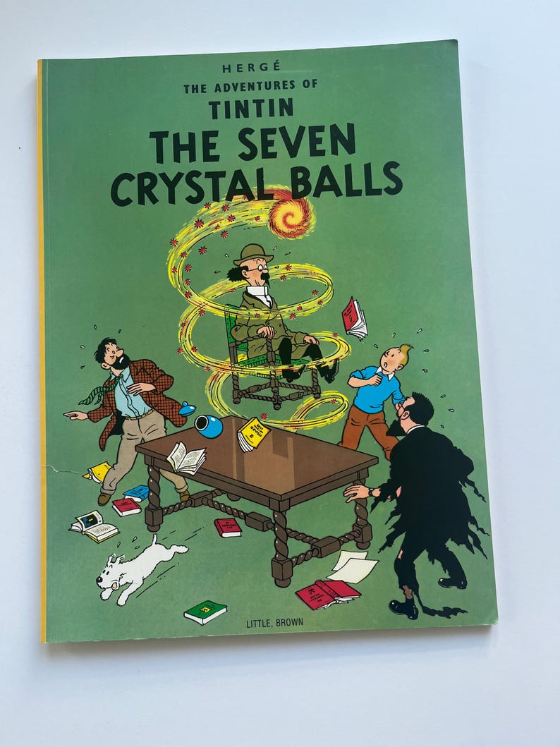 May include: A hardcover book titled "The Adventures of Tintin: The Seven Crystal Balls" with a green cover. The cover features a colorful illustration of characters and objects, including a table, books, and a dog.