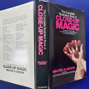 May include: A vintage book titled "The Complete Illustrated Book of Close-Up Magic" by Walter B. Gibson. The cover features a hand holding red balls. The spine displays the title in pink letters. The book is on a blue surface.